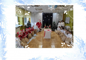 Chłopcy przebrani w mikołaje, dziewczynki za śnieżynki, siedzą na przeciwko siebie po środku wielki prezent z czerwona kokardą, z tyłu Mikołaj, Pani Zima i Pani Agnieszka.